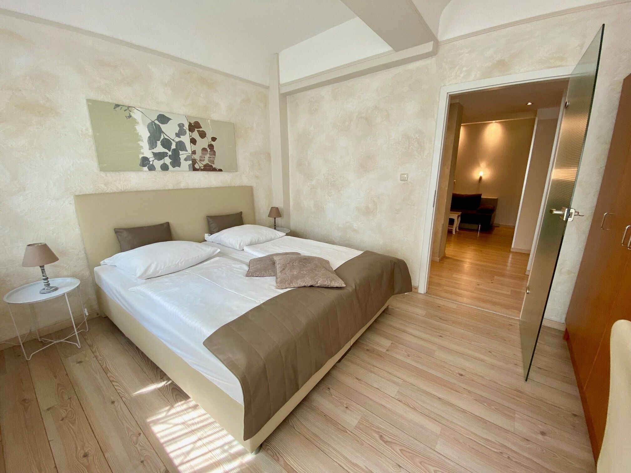 A bedroom with a bed, a nightstand, a lamp, a painting, and a door leading to another room.