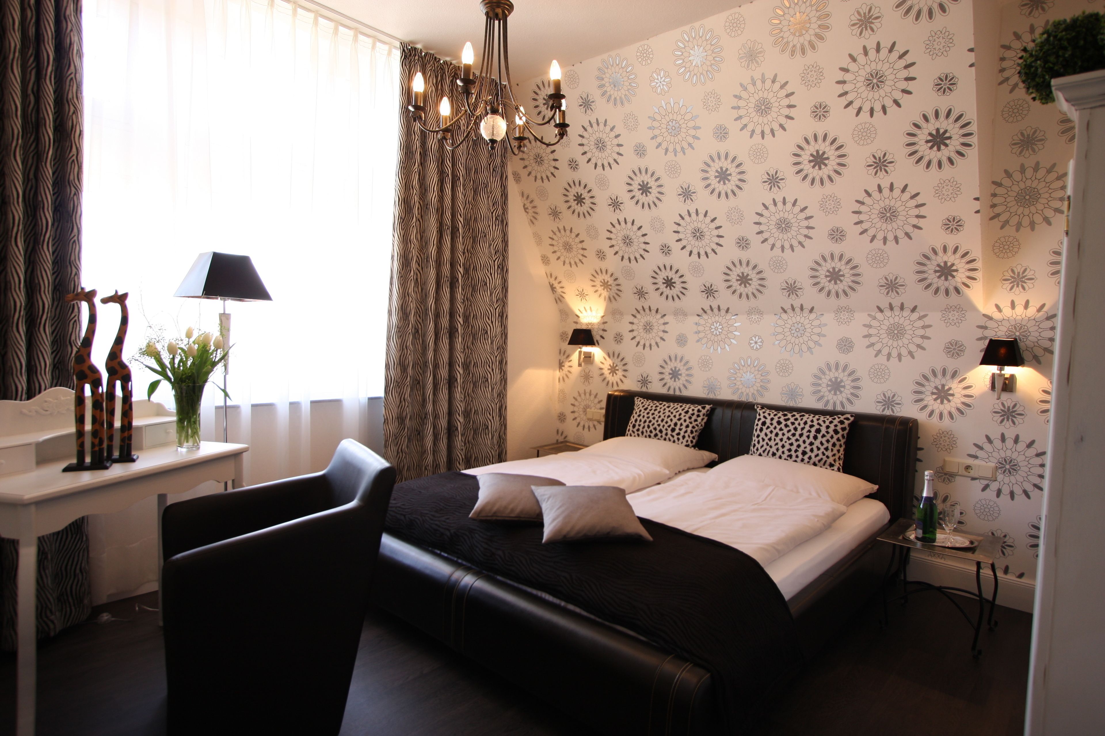 Una cama doble con cabecera oscura, una mesita de noche con lámpara y una pared con papel tapiz floral.