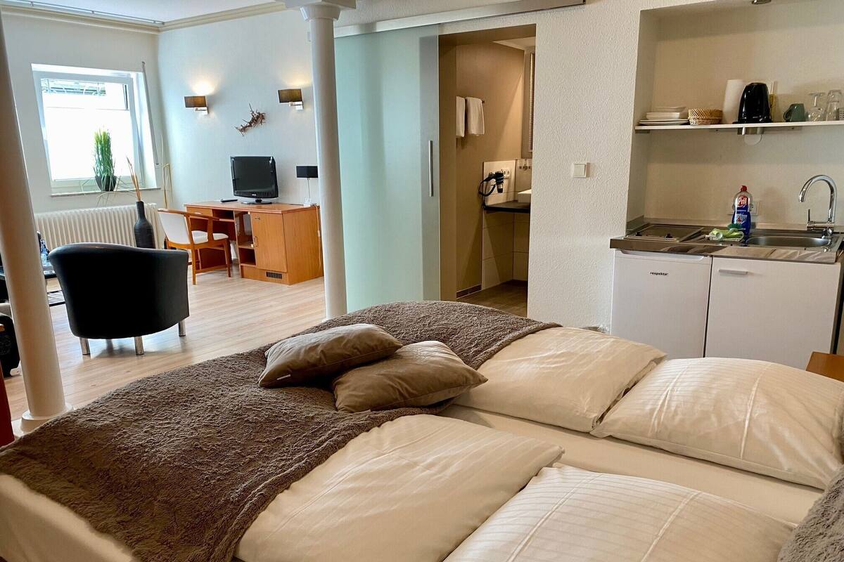 A modern hotel room with a large bed, a desk, a chair, a television, and a kitchenette.