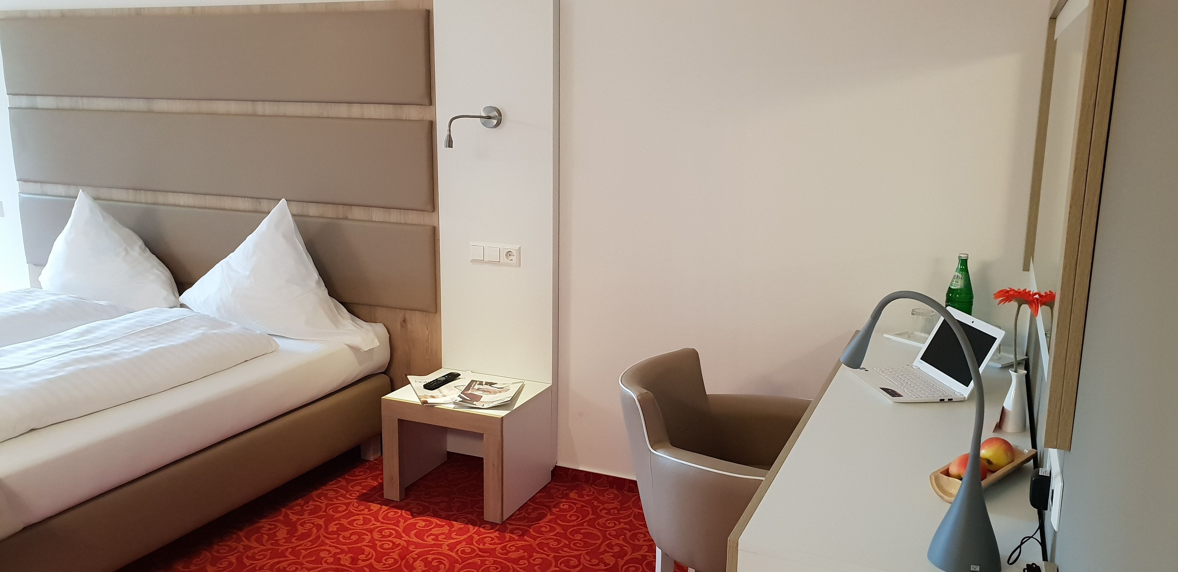 A hotel room with a bed, a small table with a remote control, a laptop, and a chair.