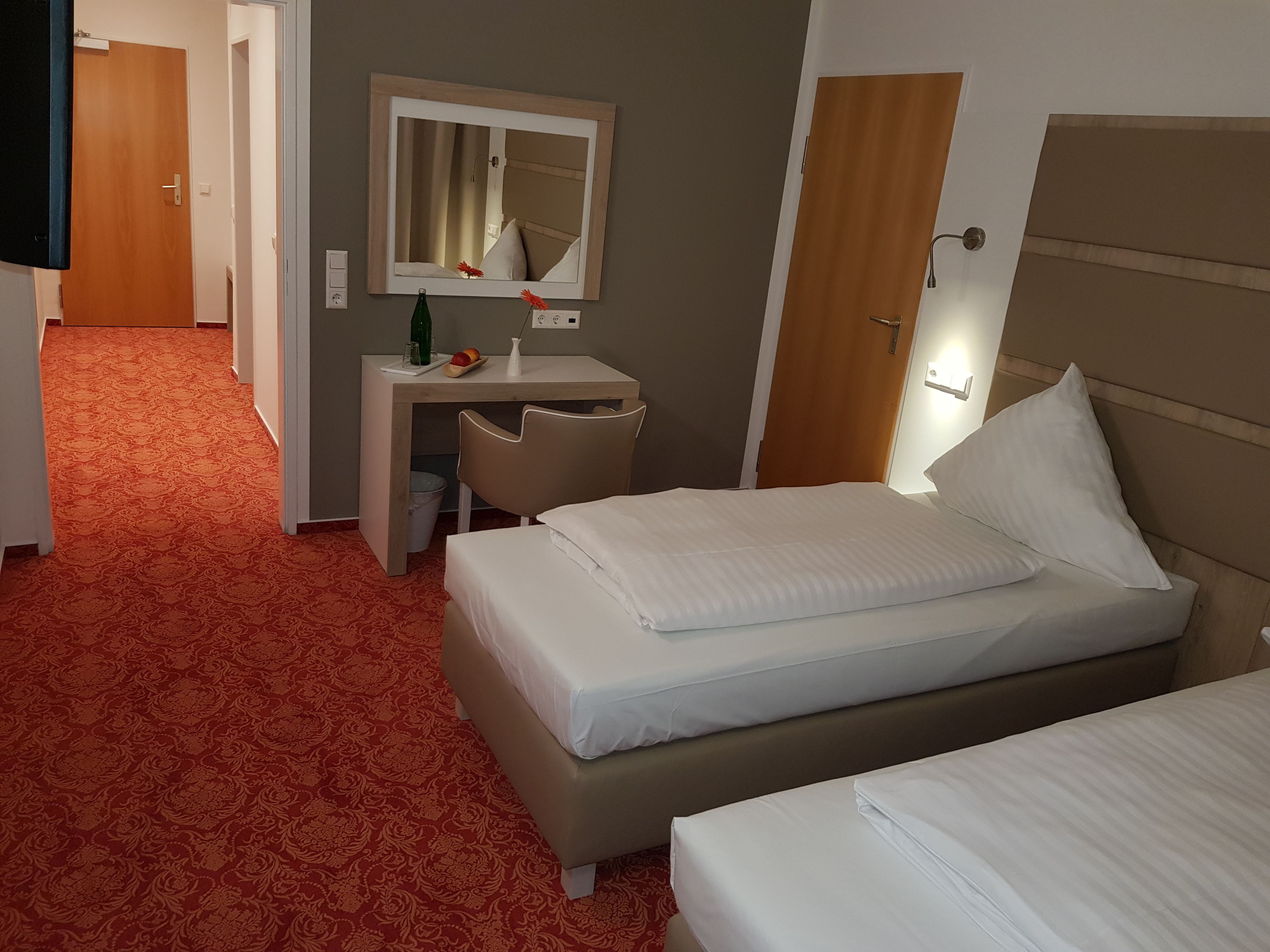 A hotel room with two beds, a desk with a mirror, a chair, and a small table with a vase and fruit.