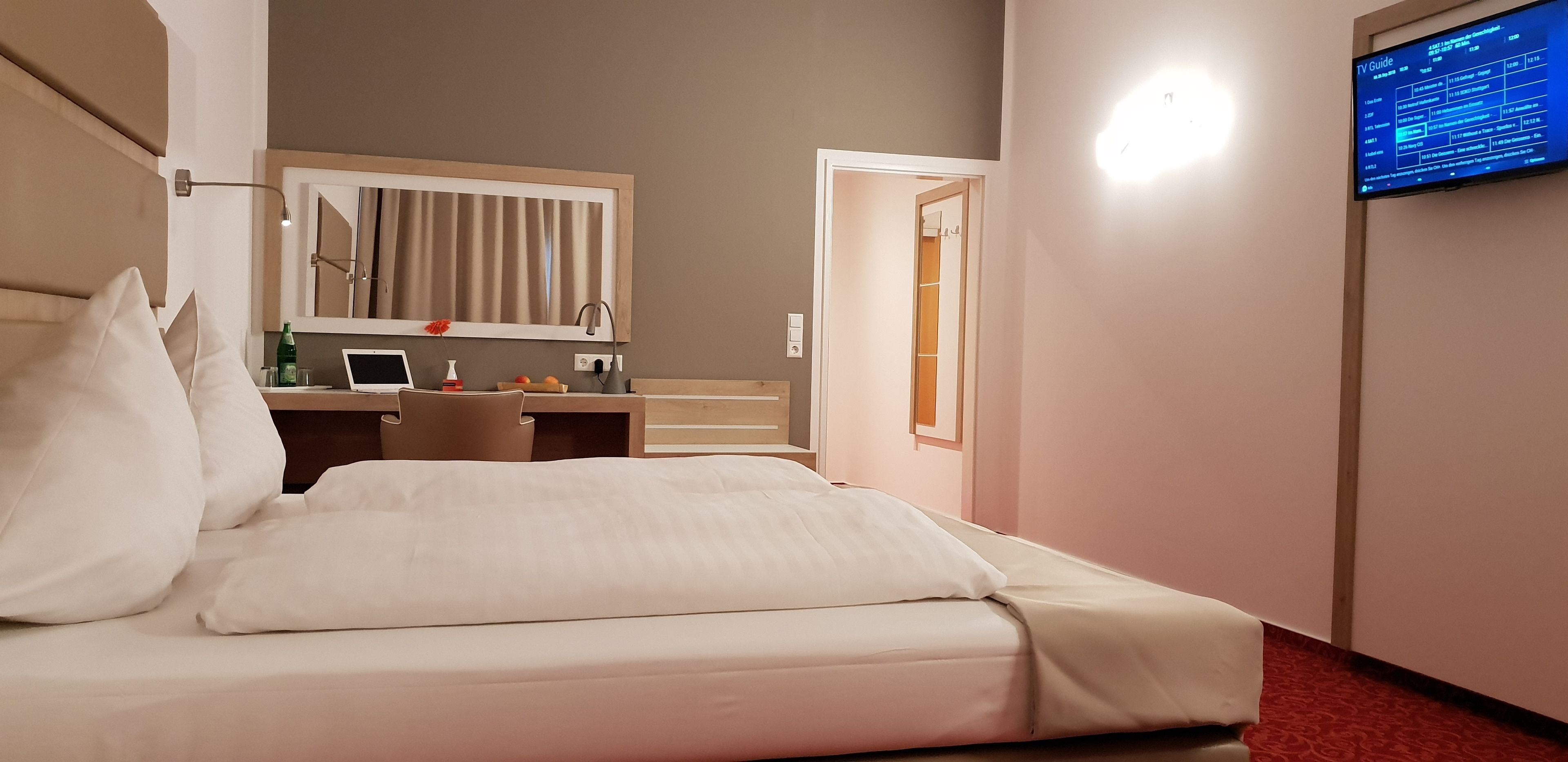 Une chambre d’hôtel équipée d’un lit, d’un bureau avec un ordinateur portable, d’une télévision affichant un programme et d’un miroir au-dessus du bureau.