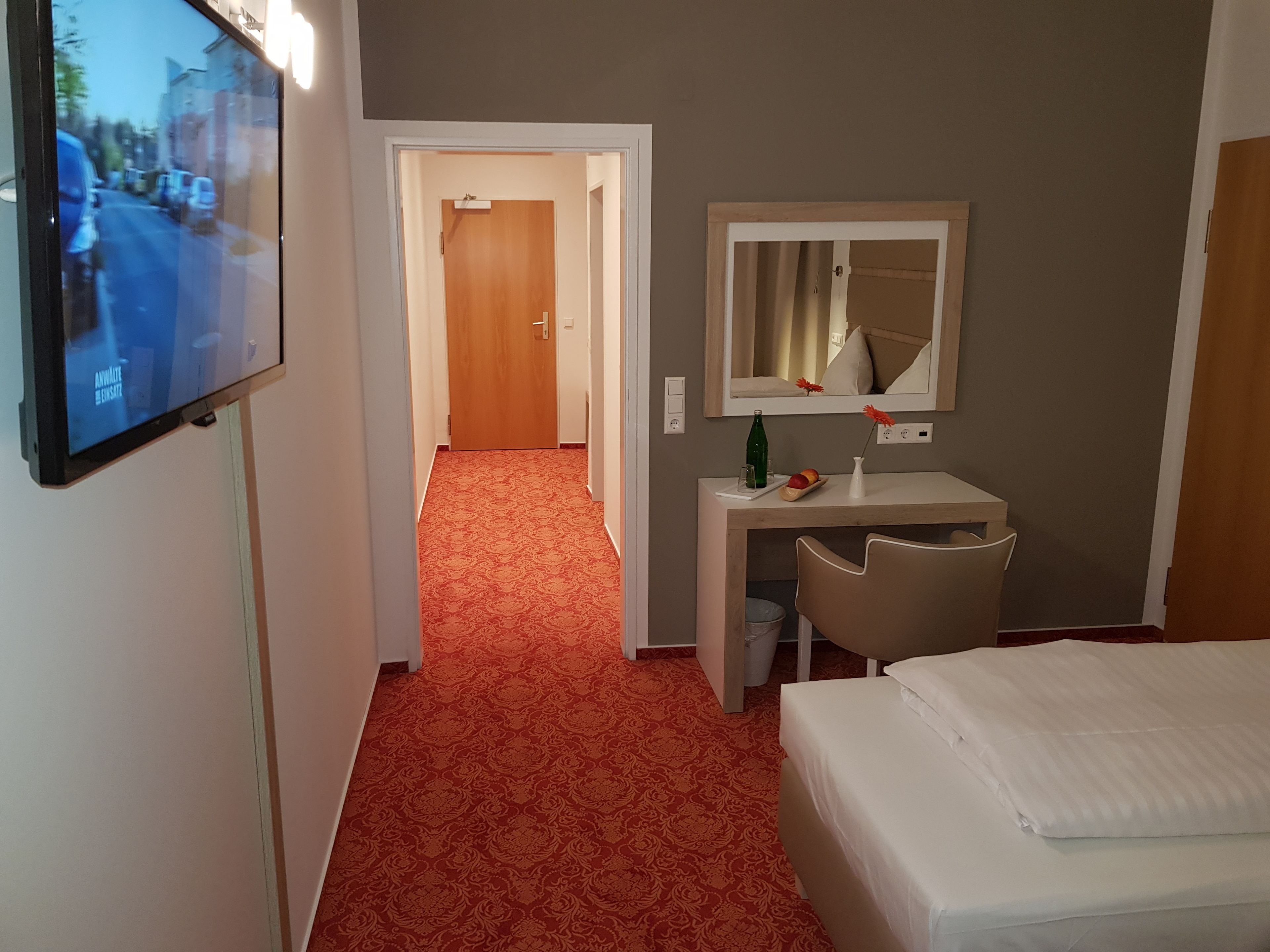 Habitación de hotel con cama, escritorio con silla, televisor montado en la pared y pasillo con alfombra.