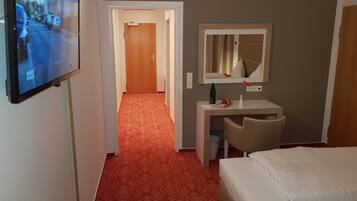 A hotel room with a bed, a desk with a chair, a TV mounted on the wall, and a carpeted hallway.
