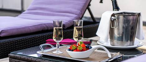 A tray with a bowl of fruit, two glasses of champagne, and a champagne bottle on a wicker table.