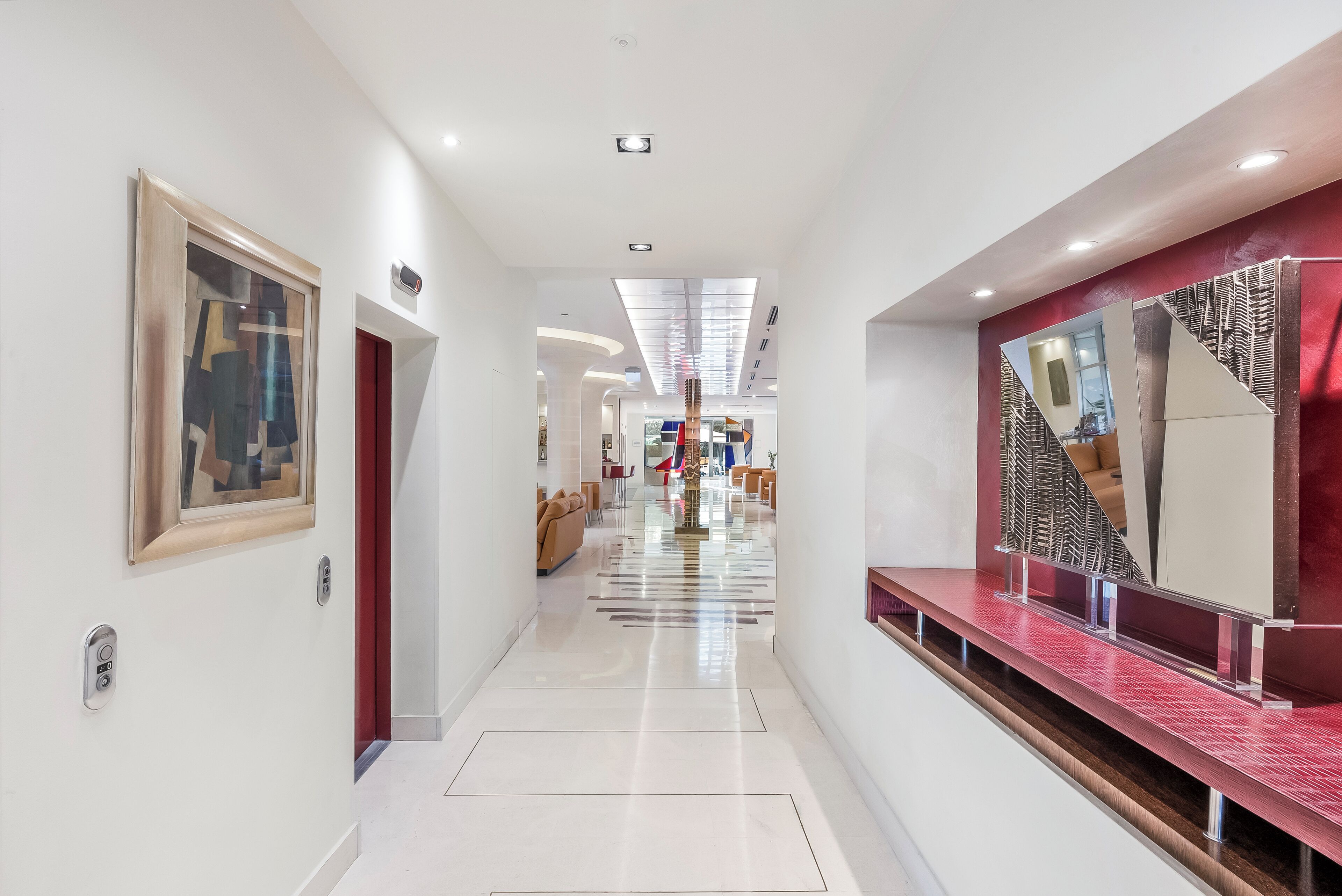A modern hotel corridor with a polished floor, a red accent wall, and a large mirror reflecting the interior.