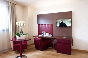 A hotel room with a desk, a television, a red chair, a red sofa, a wooden floor, and a window with curtains.