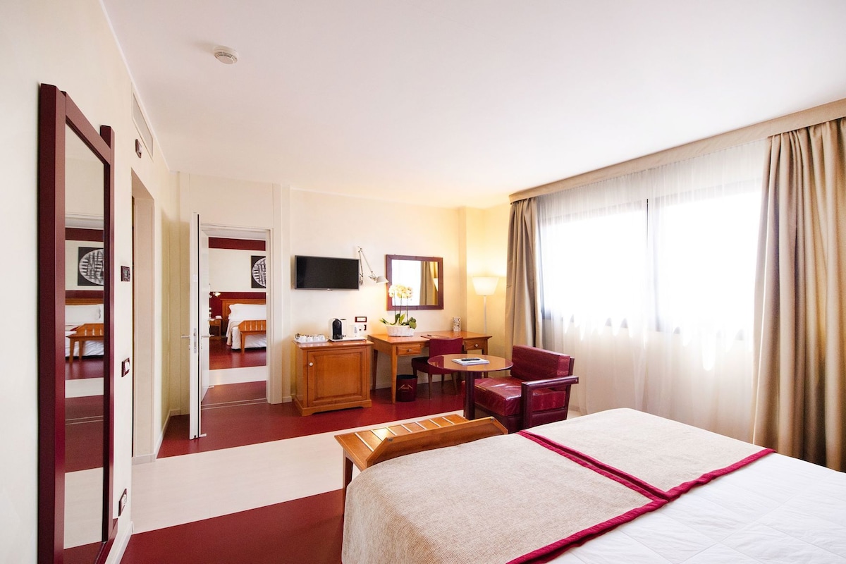 Habitación de hotel con cama, escritorio, silla, televisor y ventana con cortinas.
