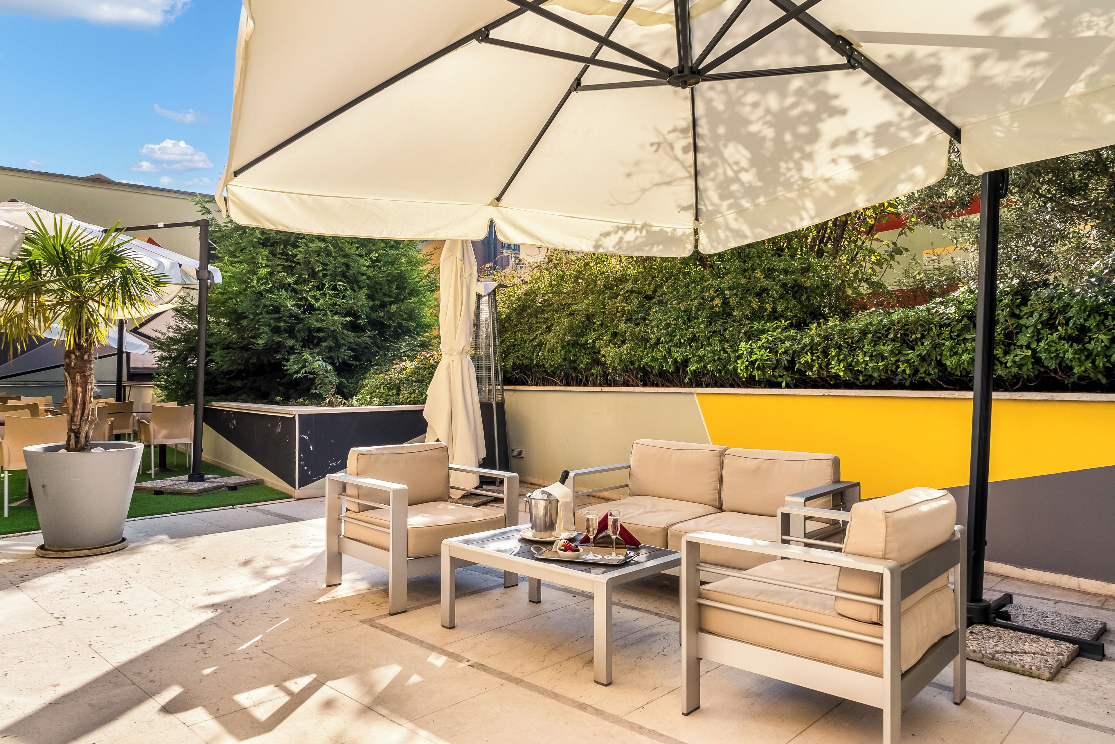An outdoor patio with a beige sofa, two armchairs, a coffee table, and a large umbrella providing shade.