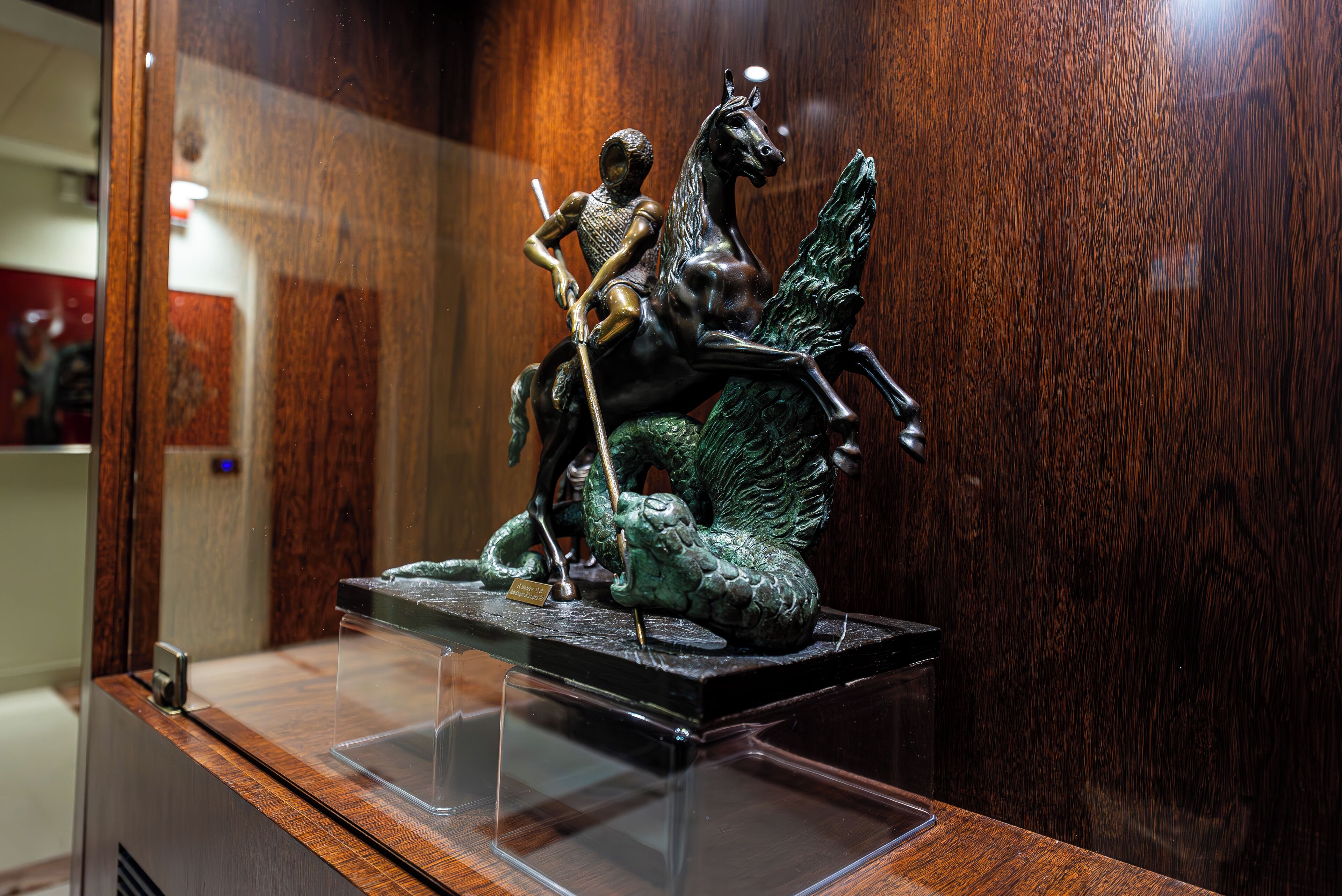 Uma estátua de bronze de uma figura montada a cavalo, exposta sobre um pedestal de madeira numa vitrine de vidro.