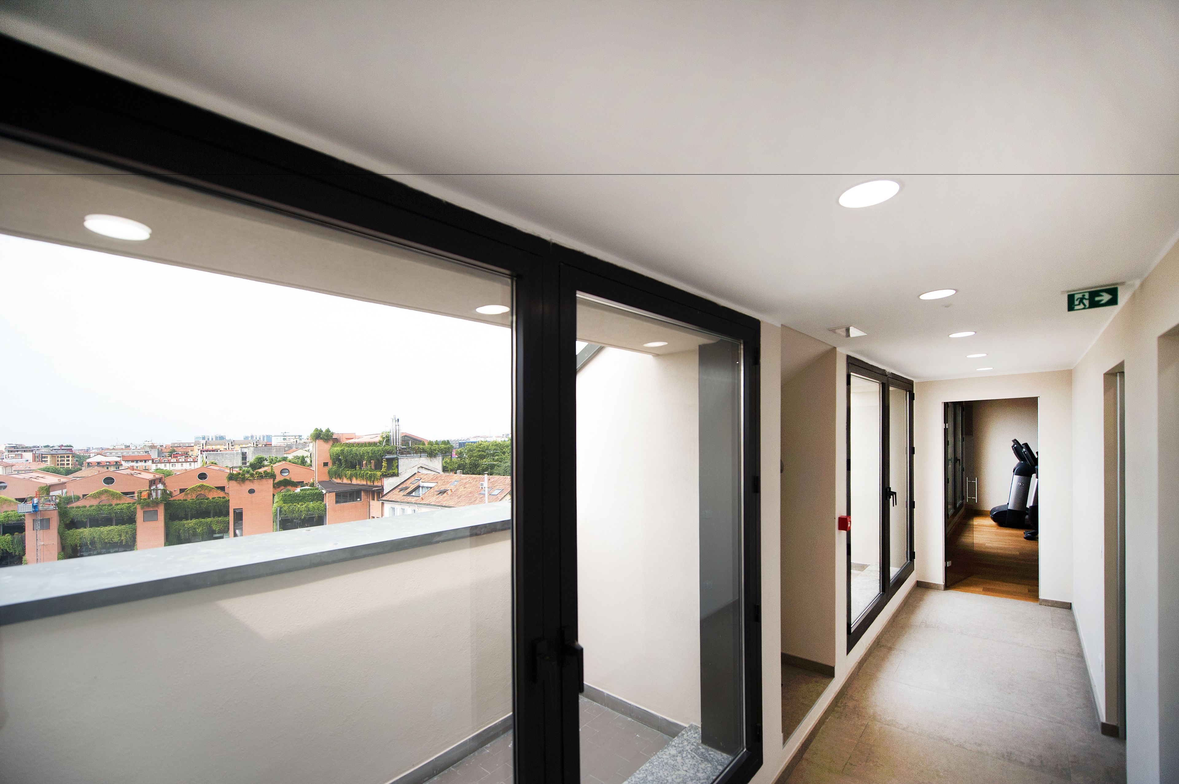 Un pasillo con vistas a edificios exteriores, una cinta de correr a lo lejos y una puerta con letrero de salida de emergencia.