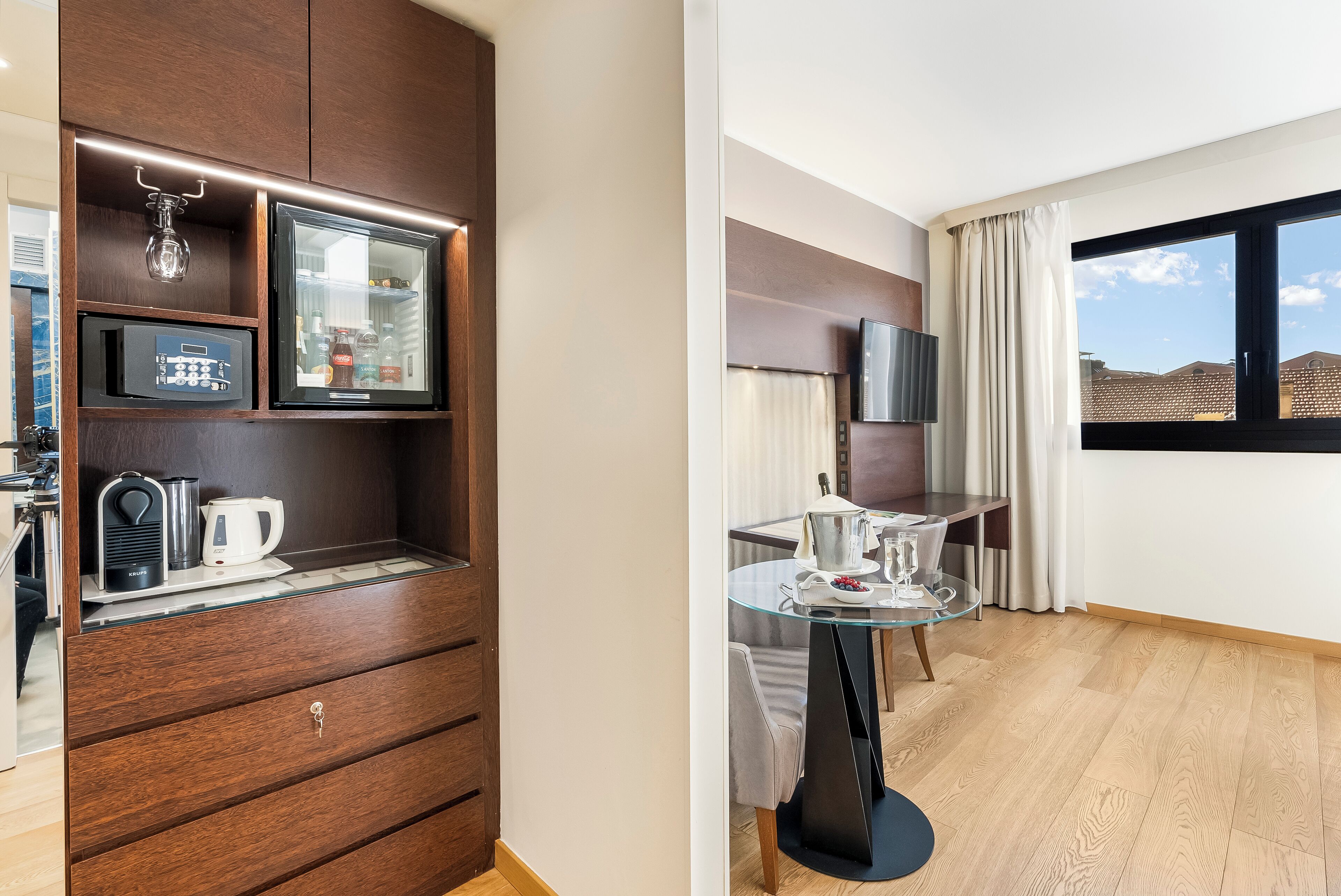 A modern hotel room with a wooden wardrobe, a glass-top dining table, a flat-screen TV, and a large window with a view.