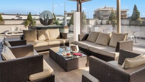 Un área de estar al aire libre iluminada por el sol, con mobiliario de mimbre y una mesa de centro de vidrio, con vistas a un paisaje urbano.
