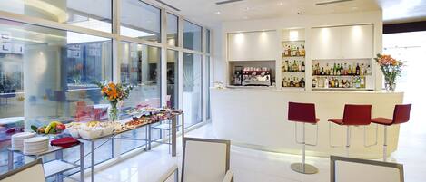 Un moderno vestíbulo de hotel con un separador de vidrio, una zona de bar con taburetes y una mesa de buffet con comida y bebidas.
