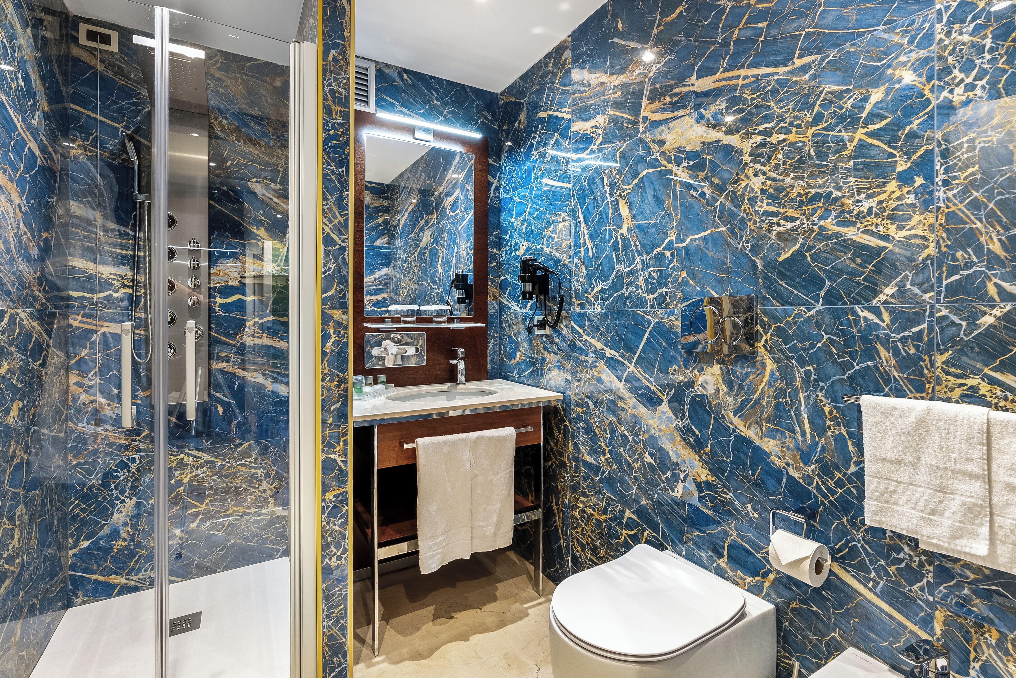 A bathroom with a marble wall, a glass-enclosed shower, a sink with a mirror, and a toilet.