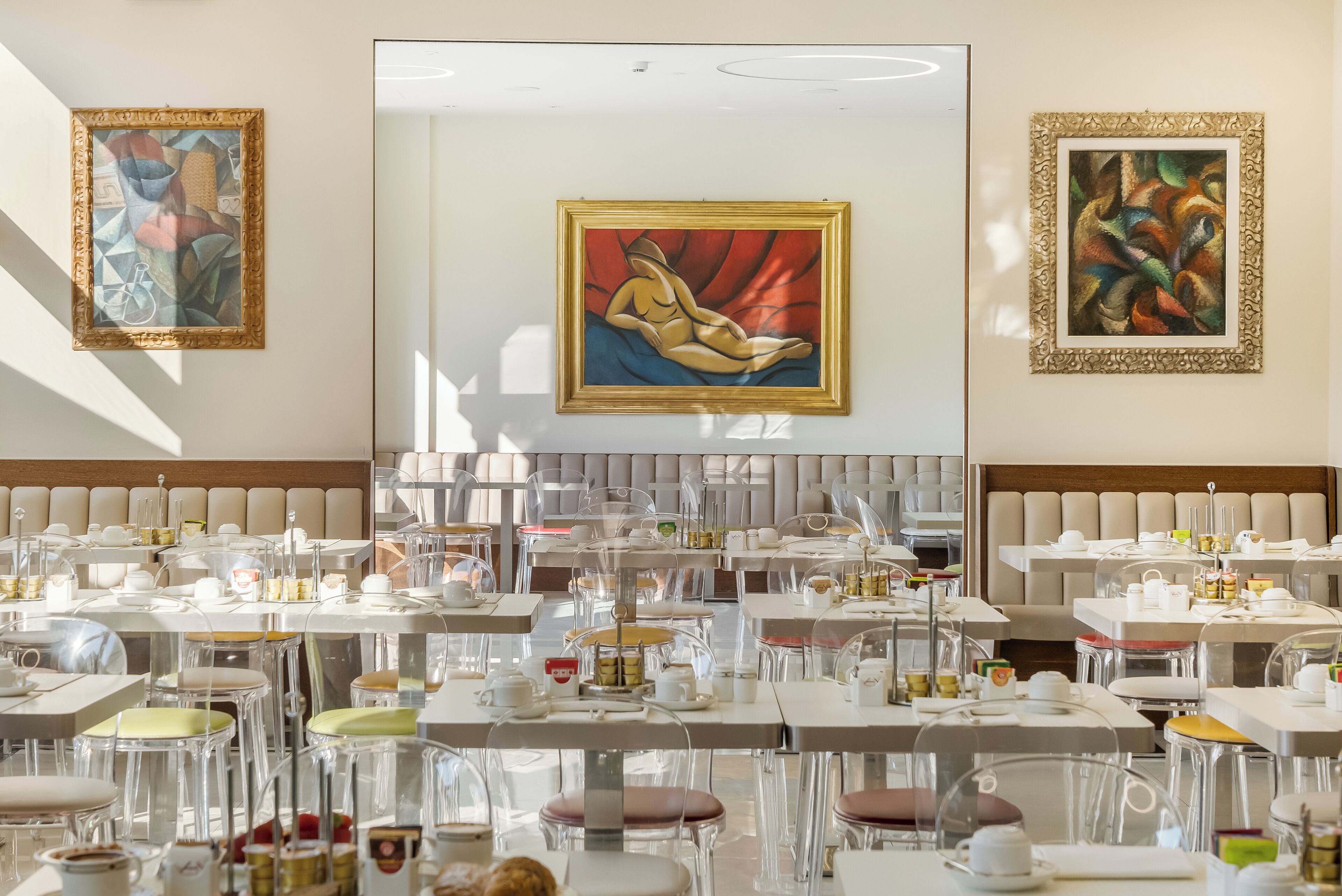A modern dining area with clear glass tables and chairs, framed artwork on the walls, and a neutral color palette.