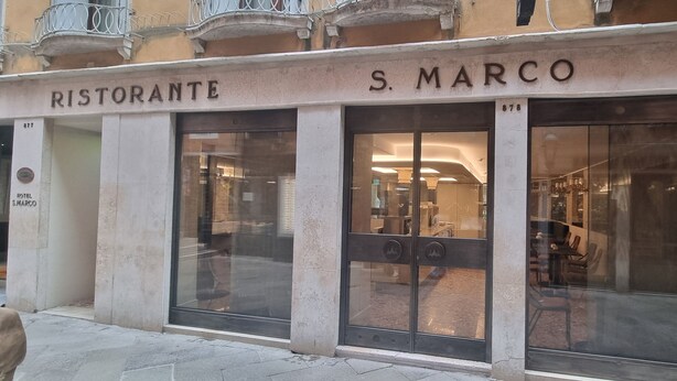 Exterior de un restaurante llamado "Ristorante S. Marco", con amplios ventanales de vidrio y fachada de piedra.