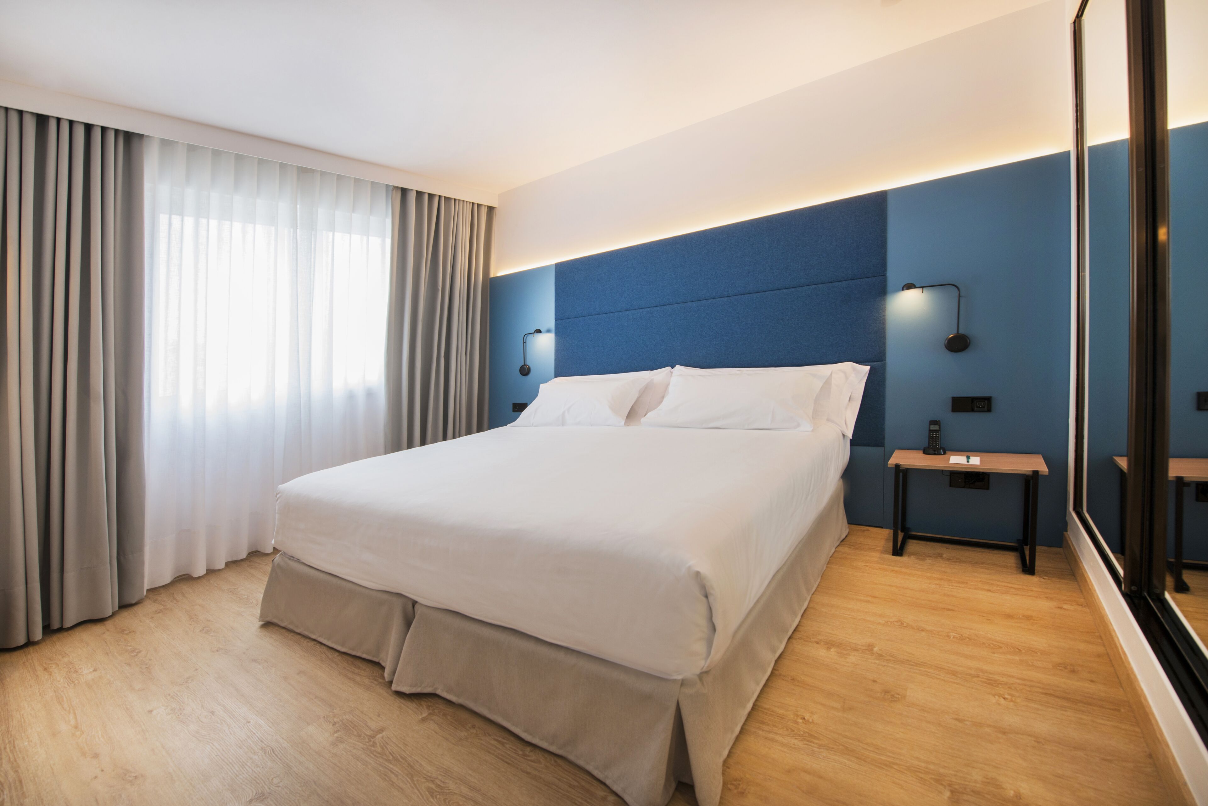 Una habitación de hotel moderna con una cama grande, suelo de madera, una pared azul y una mesita de noche con lámpara.