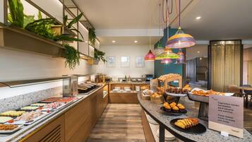 Un bufé de desayuno con diversos alimentos, un letrero que indica productos horneados recién hechos, y un diseño interior moderno con detalles de madera y lámparas colgantes.