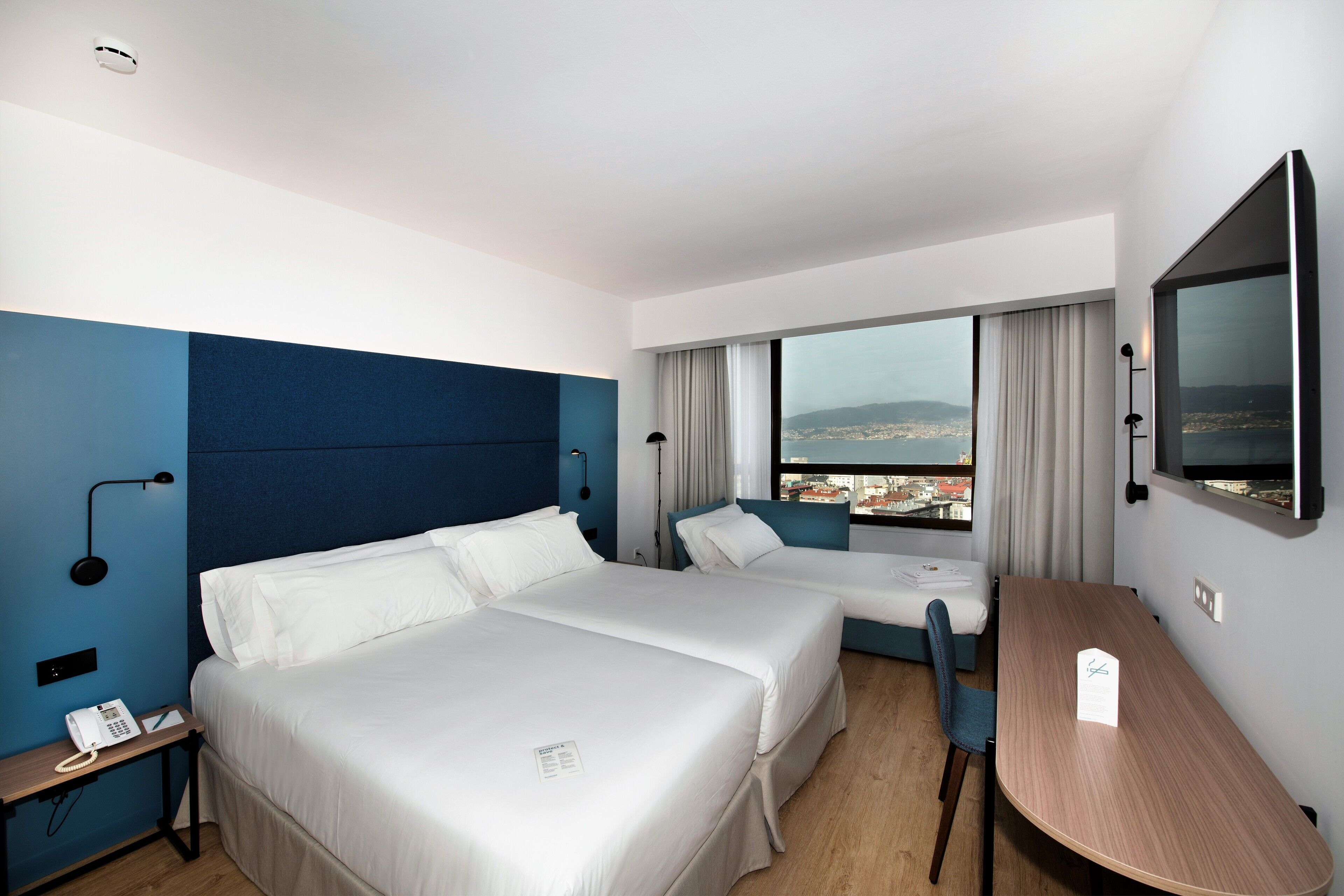 Habitación de hotel con dos camas, un escritorio, una silla, un televisor y ventana con vistas a la ciudad.