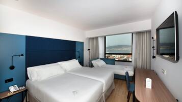 Habitación de hotel con dos camas, un escritorio, una silla, un televisor y ventana con vistas a la ciudad.