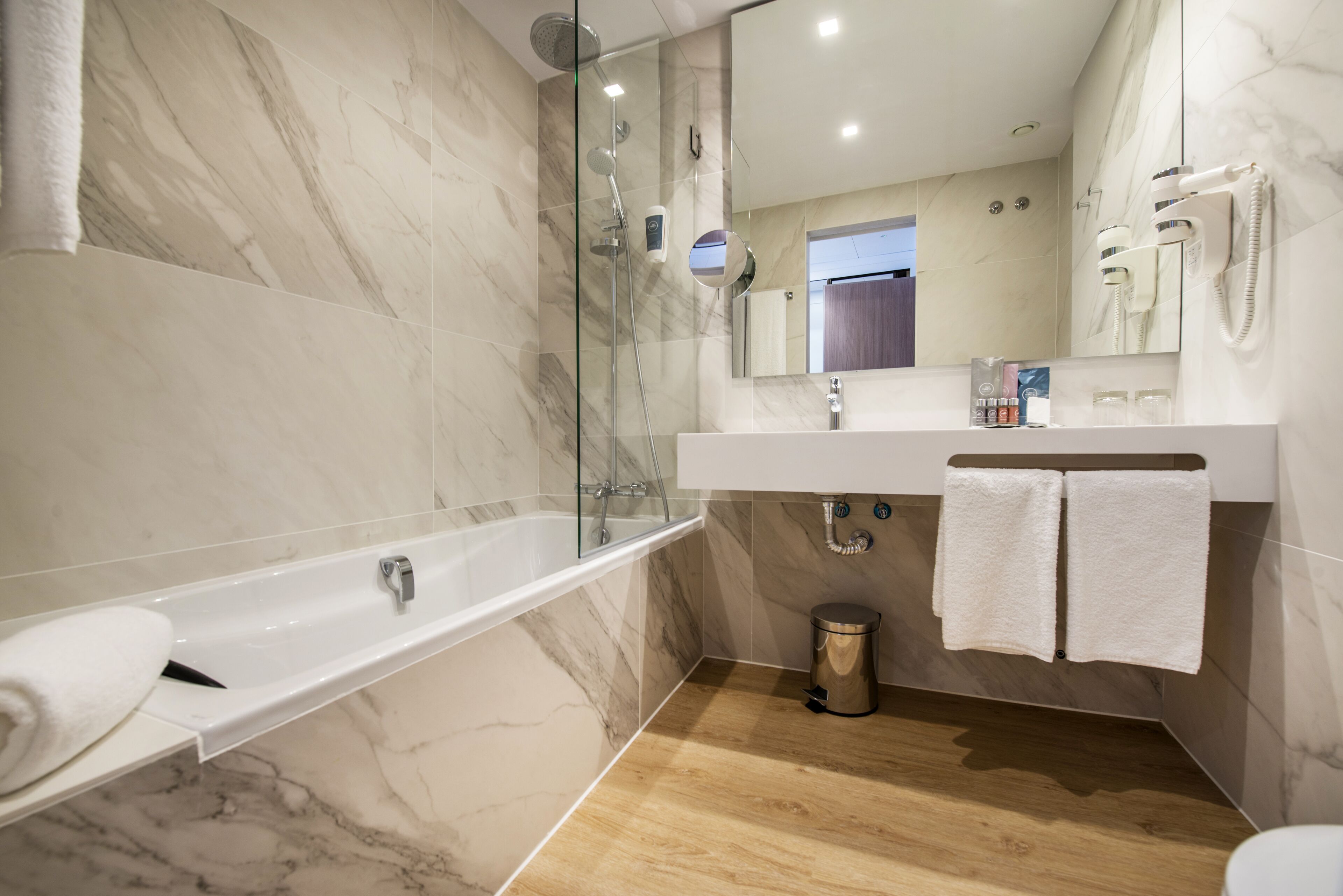 A modern bathroom with a glass-enclosed shower, a white bathtub, a vanity with a mirror, and a wooden floor.