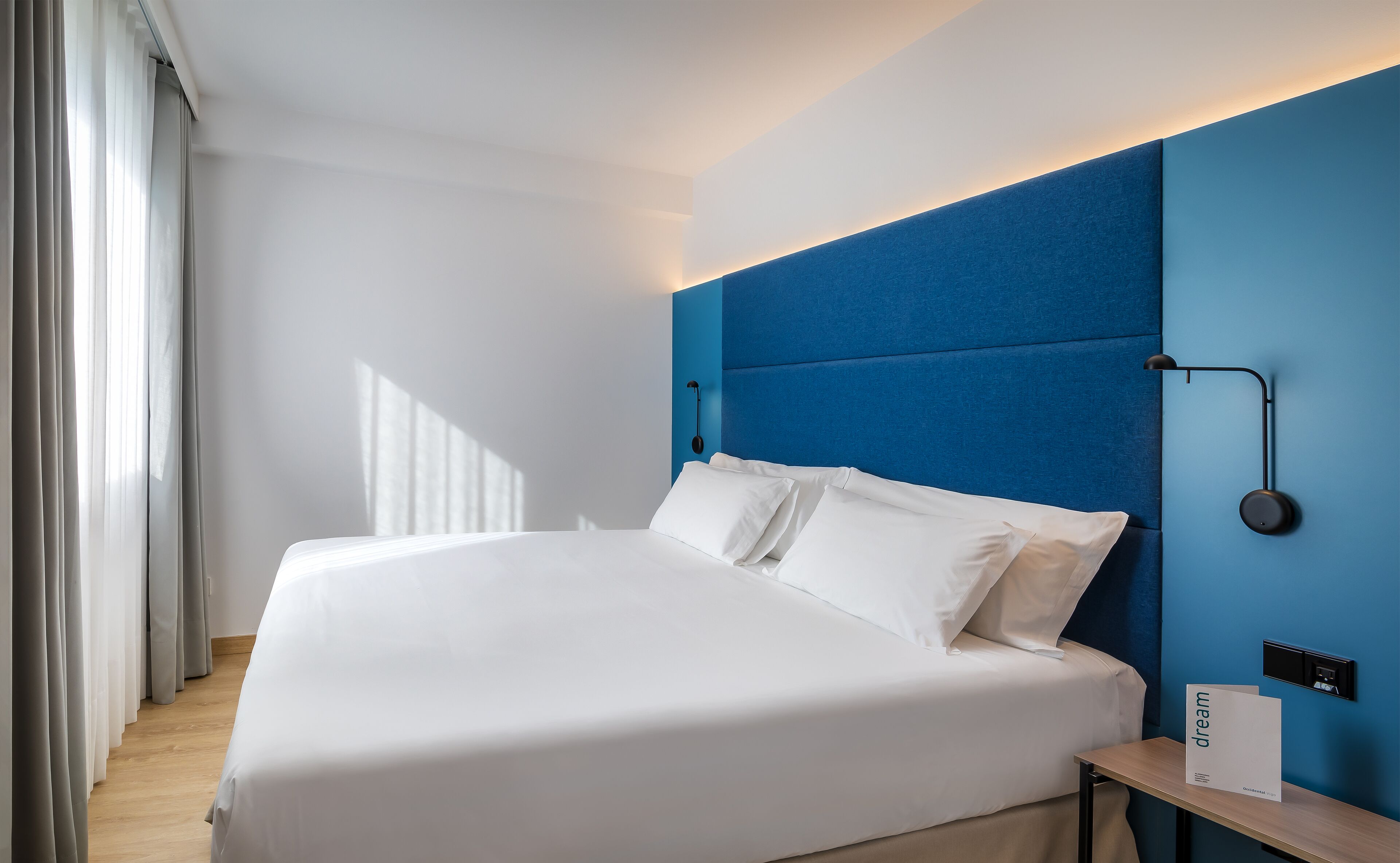 A modern hotel room with a large bed, a bedside table, and a wall-mounted lamp.