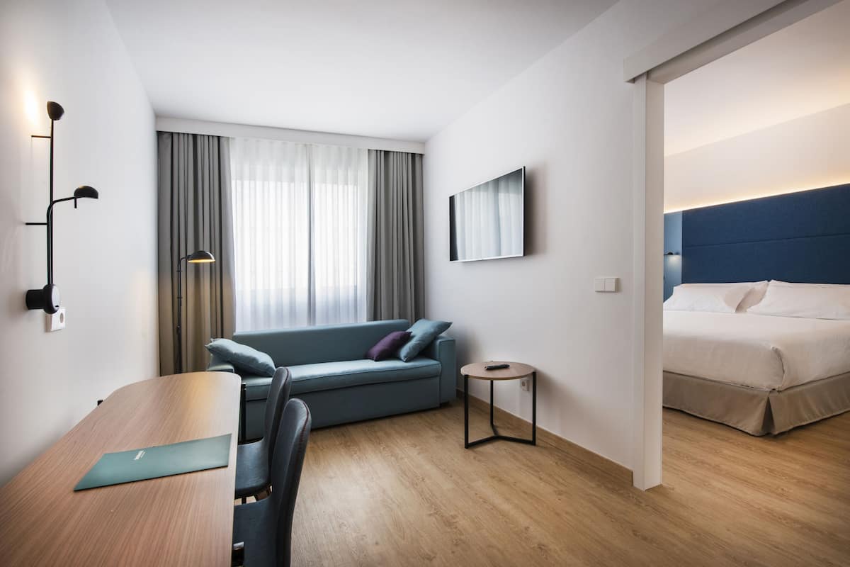 Ett modernt hotellrum med trägolv, en soffa, en säng, ett litet runt bord och ett skrivbord med en bärbar dator.