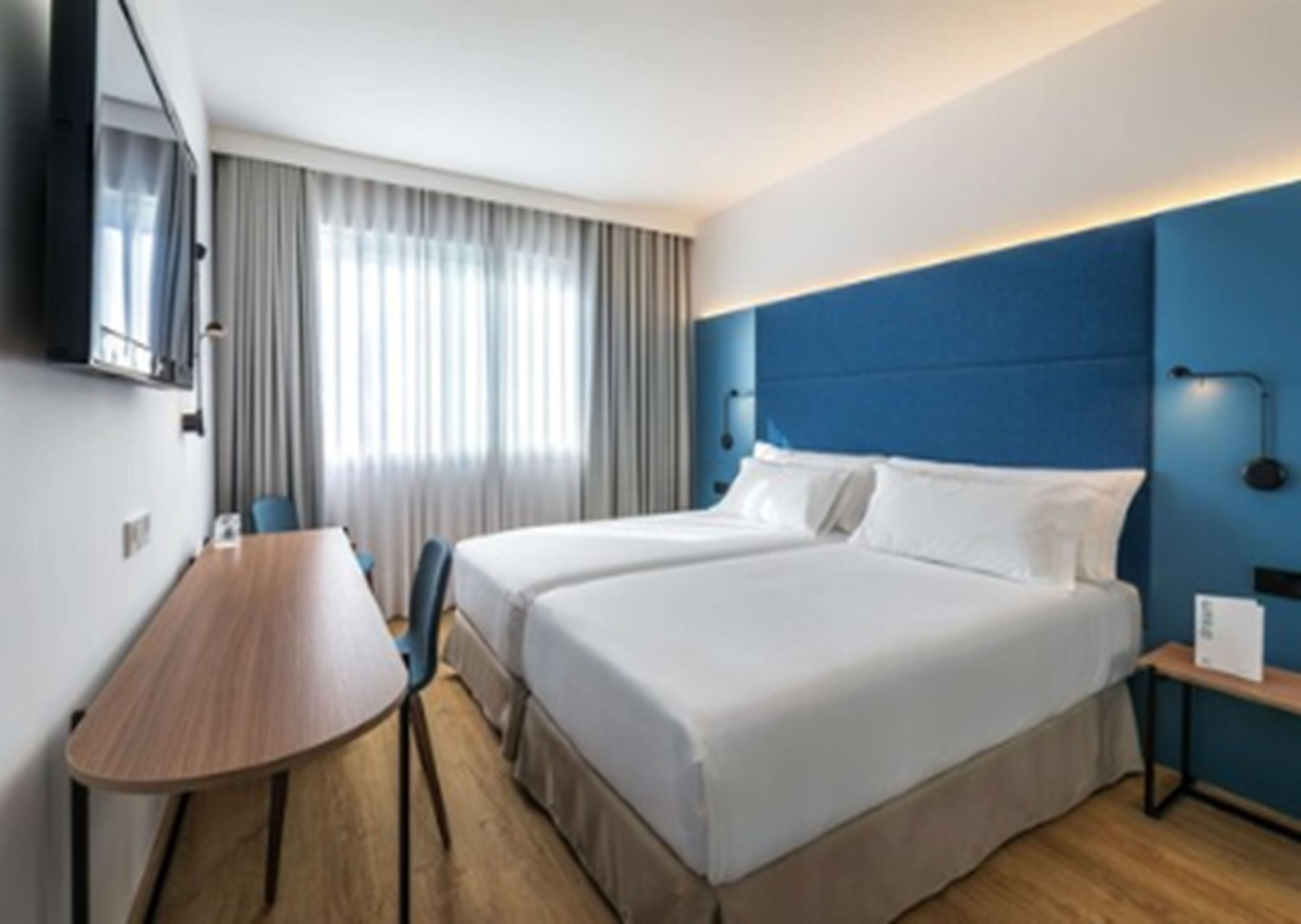 A hotel room with a large bed, a desk, a chair, a TV, and a window with curtains.