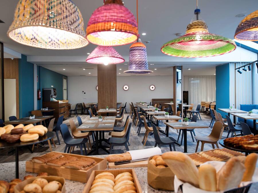 A modern dining area with multiple tables and chairs, colorful hanging lamps, and a variety of bread displayed on tables.