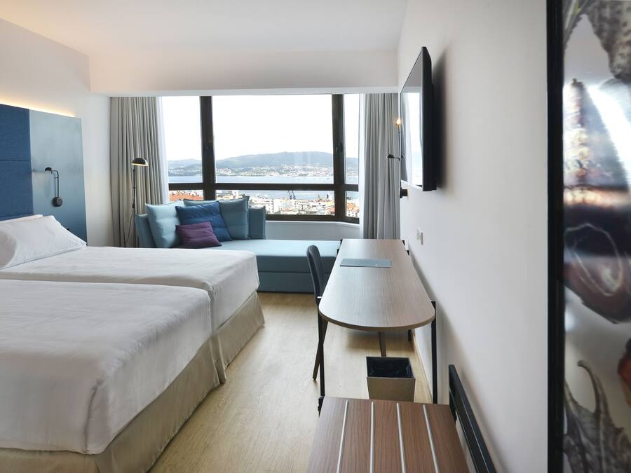 A hotel room with two beds, a desk, a chair, a view of the city, and a TV on the wall.