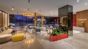 A modern hotel lobby with a seating area, a reception desk, and a large window overlooking a parking area. - Occidental Vigo (Vigo)