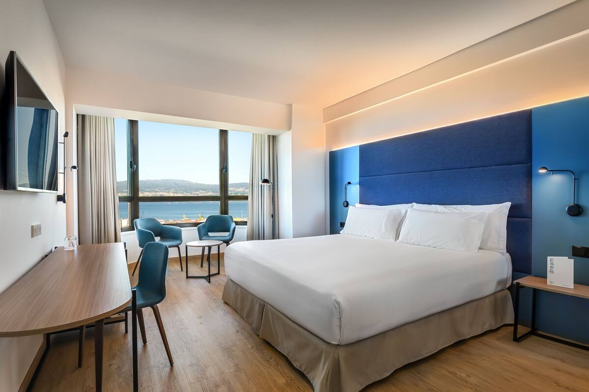 Ett modernt hotellrum med en stor säng, ett skrivbord och en stol. Genom de stora fönstren har man utsikt över havet.
