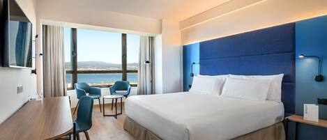 Habitación de hotel moderna con una cama grande, un escritorio y una silla. Se ve el mar a través de ventanas amplias.