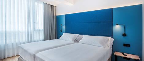 Habitación de hotel con dos camas, un muro azul decorativo y una mesita de noche con un control remoto.