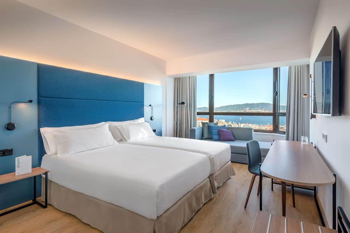 Ett modernt hotellrum med två sängar, ett matbord och ett stort fönster med utsikt över staden.