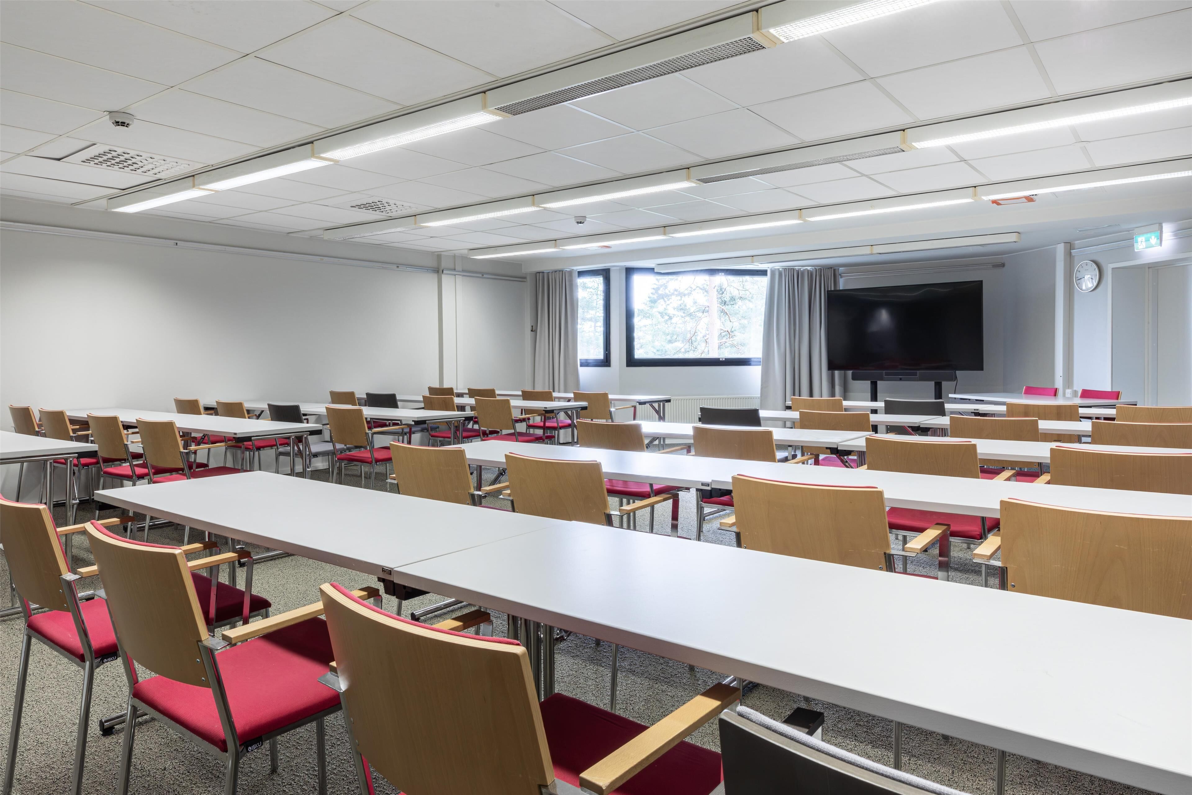 A conference room with multiple tables and chairs, a large screen, and windows with curtains.