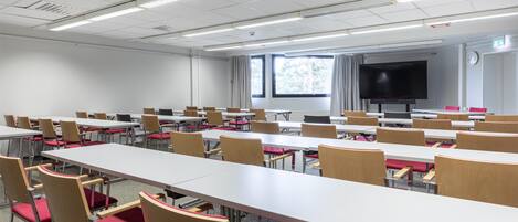 A conference room with multiple tables and chairs, a large screen, and windows with curtains.