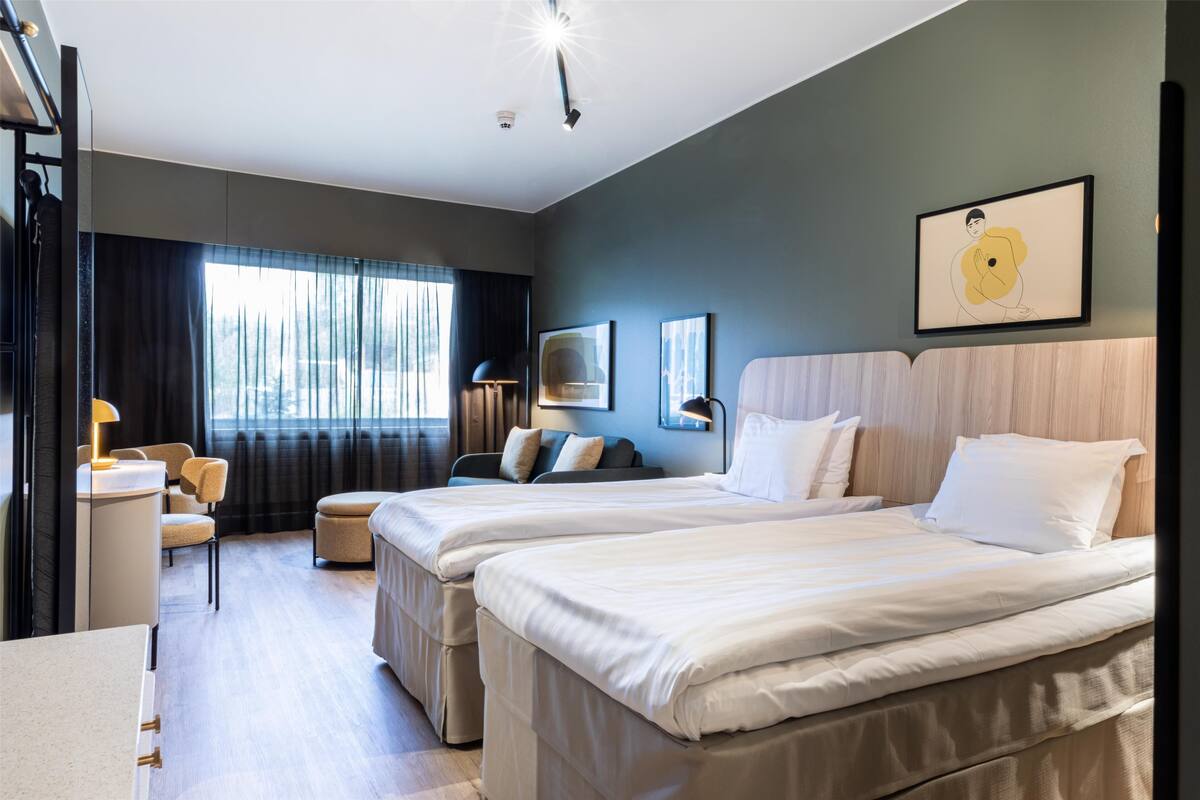 Une chambre d’hôtel moderne avec deux lits, un bureau et une chaise. On y trouve une grande fenêtre avec des rideaux, un luminaire au plafond et un tableau encadré au mur.