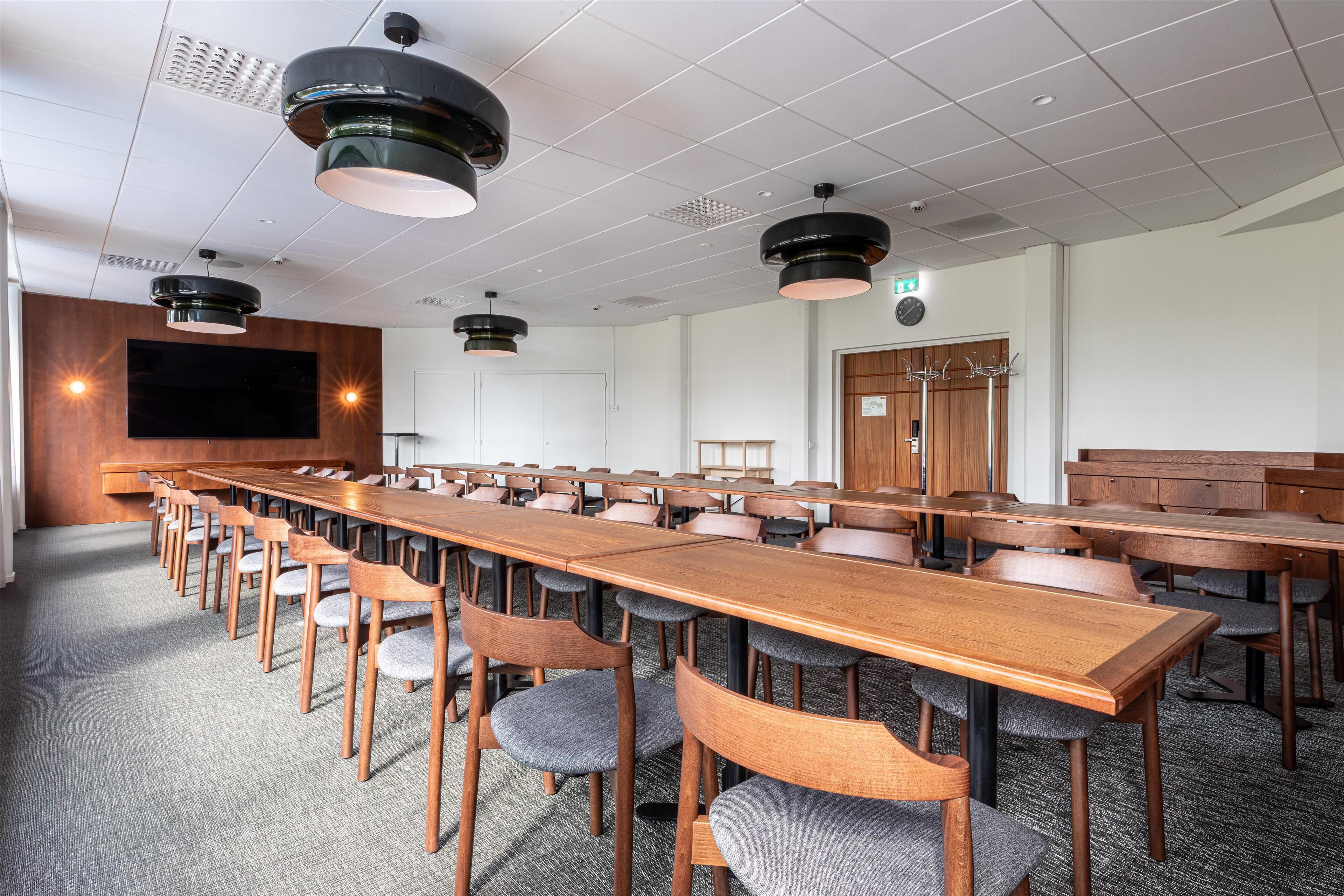 A conference room with long tables and chairs, ceiling lights, a large screen, and a wooden door.