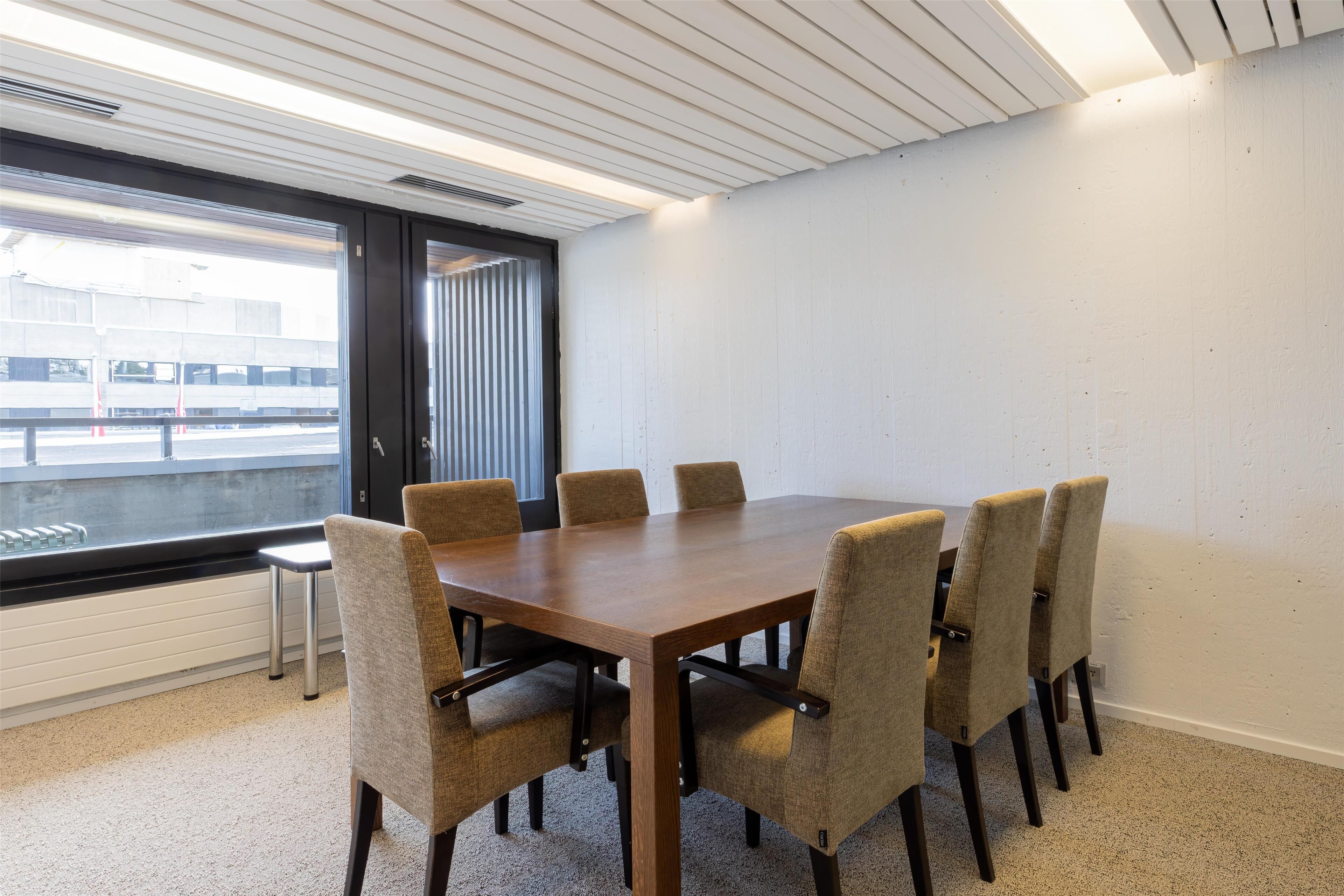 A conference room with a large table and six chairs, a window with blinds, and a white wall.