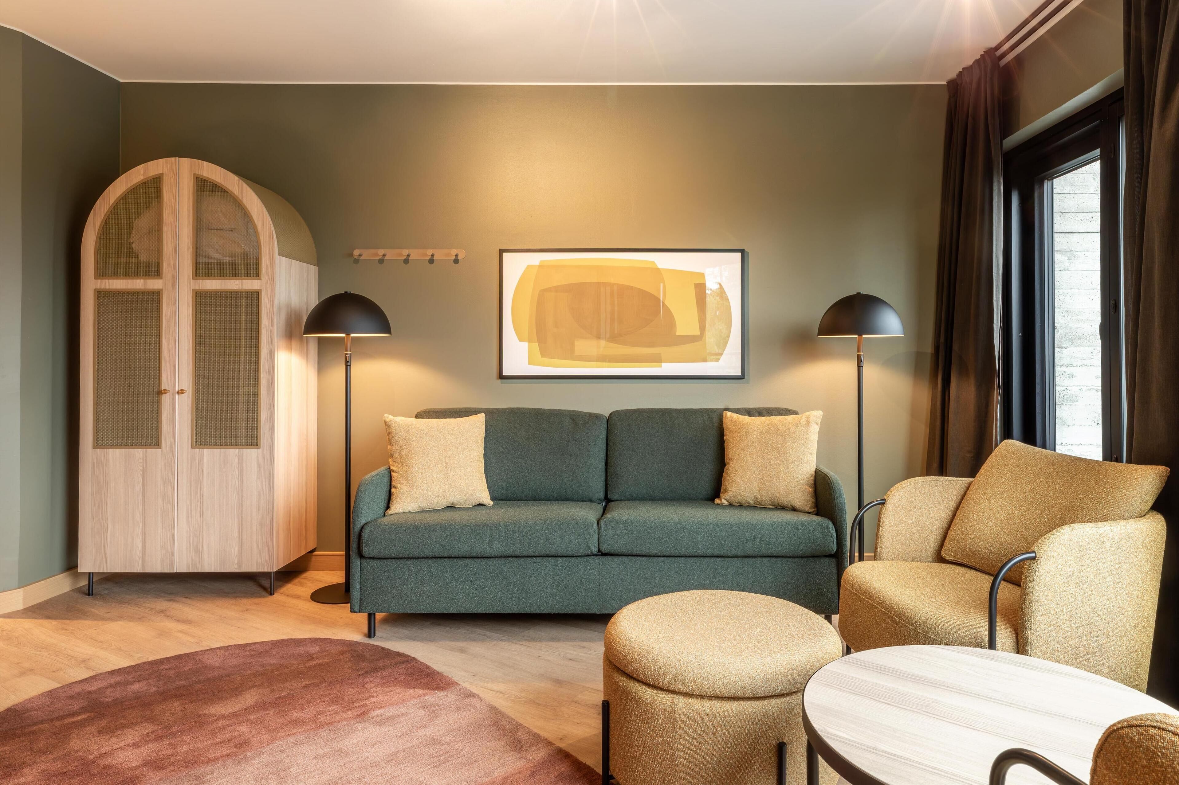 A modern living room with a green sofa, a beige armchair, a round ottoman, and a floor lamp.