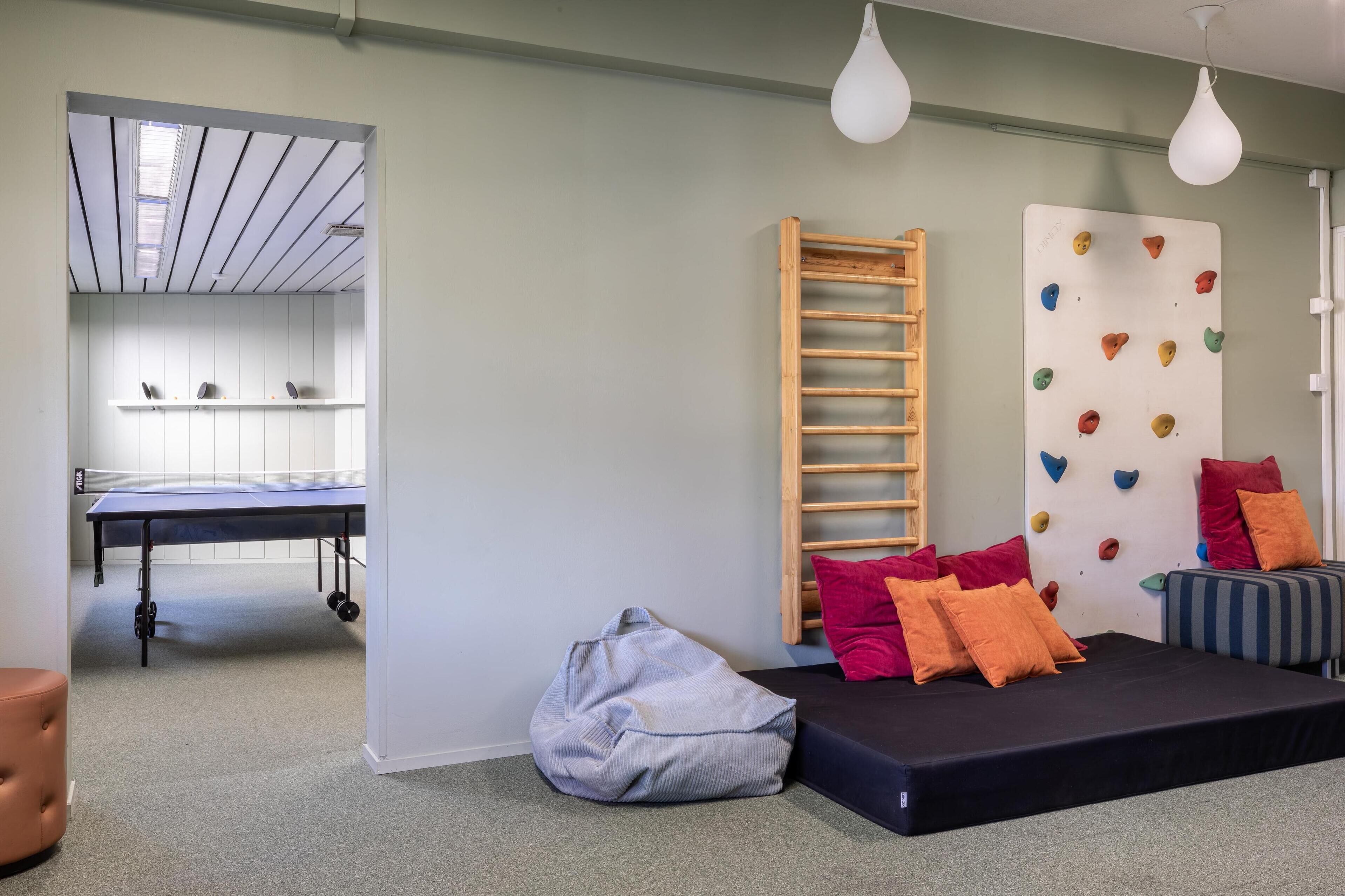 A room with a ping pong table, climbing wall, bean bag, and a sofa with cushions.