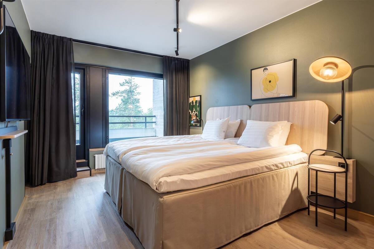 Une chambre à coucher moderne avec un grand lit, une table de chevet, une lampe sur pied et une fenêtre avec des rideaux.