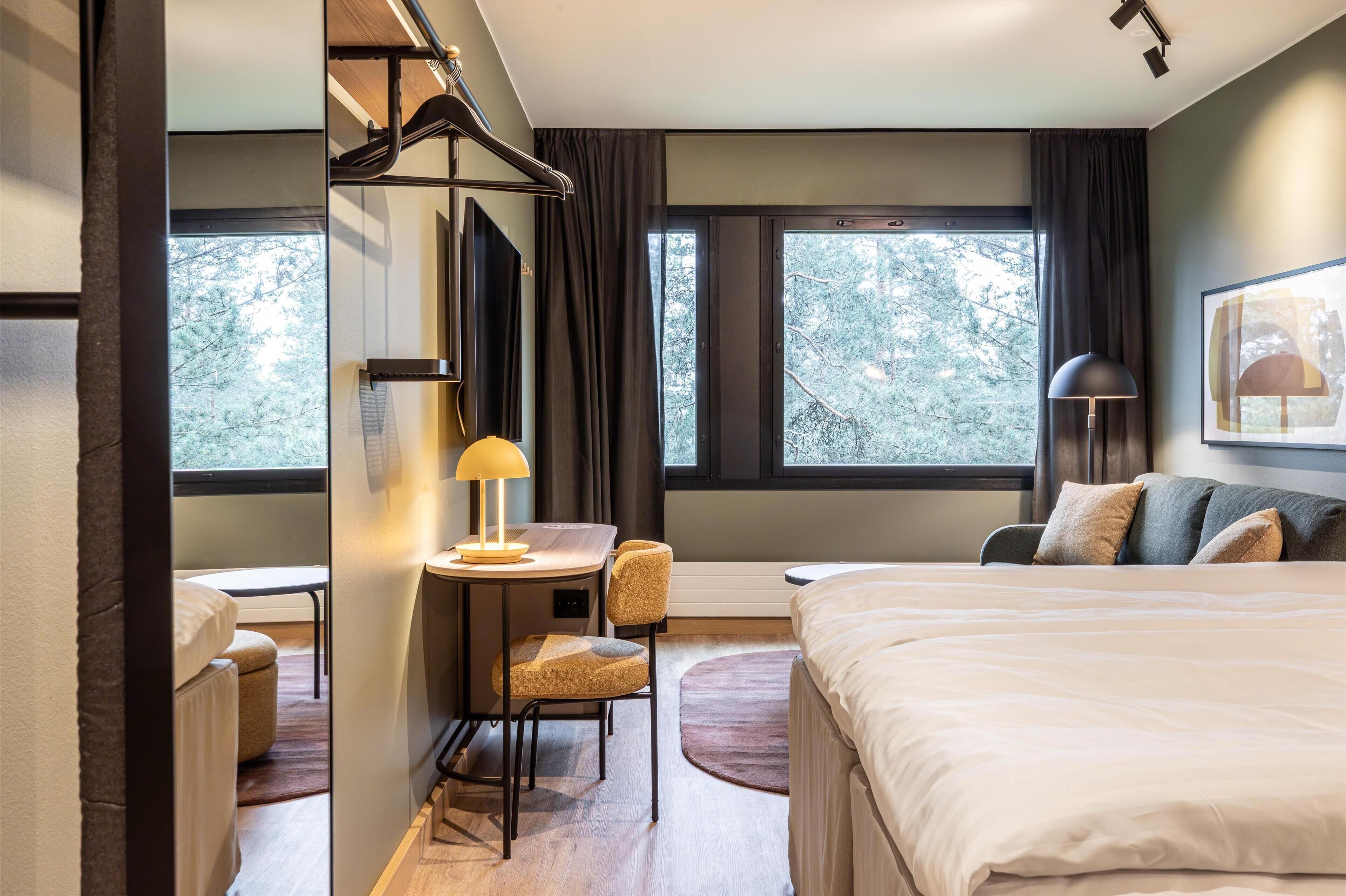 Habitación de hotel con cama, escritorio con silla, una mesita de noche con lámpara y ventanas amplias con vista a árboles.