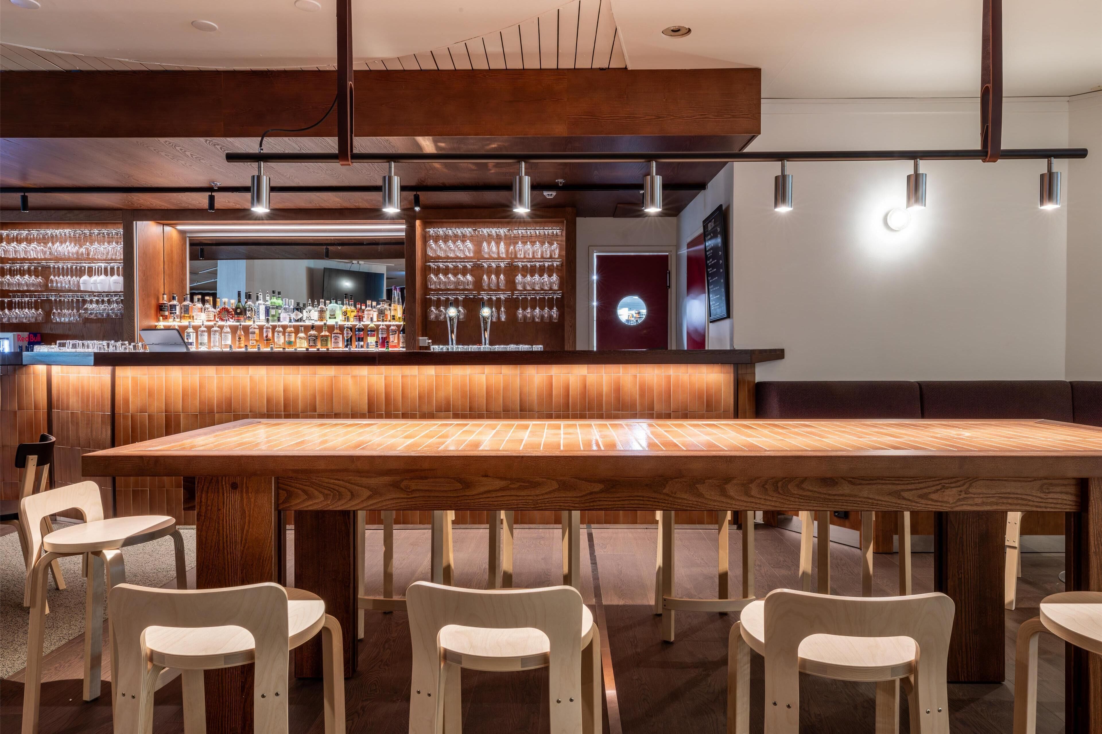 Uma área de bar com uma longa mesa de madeira e várias cadeiras, um balcão de bar com garrafas e copos, e uma parede com prateleiras cheias de copos.