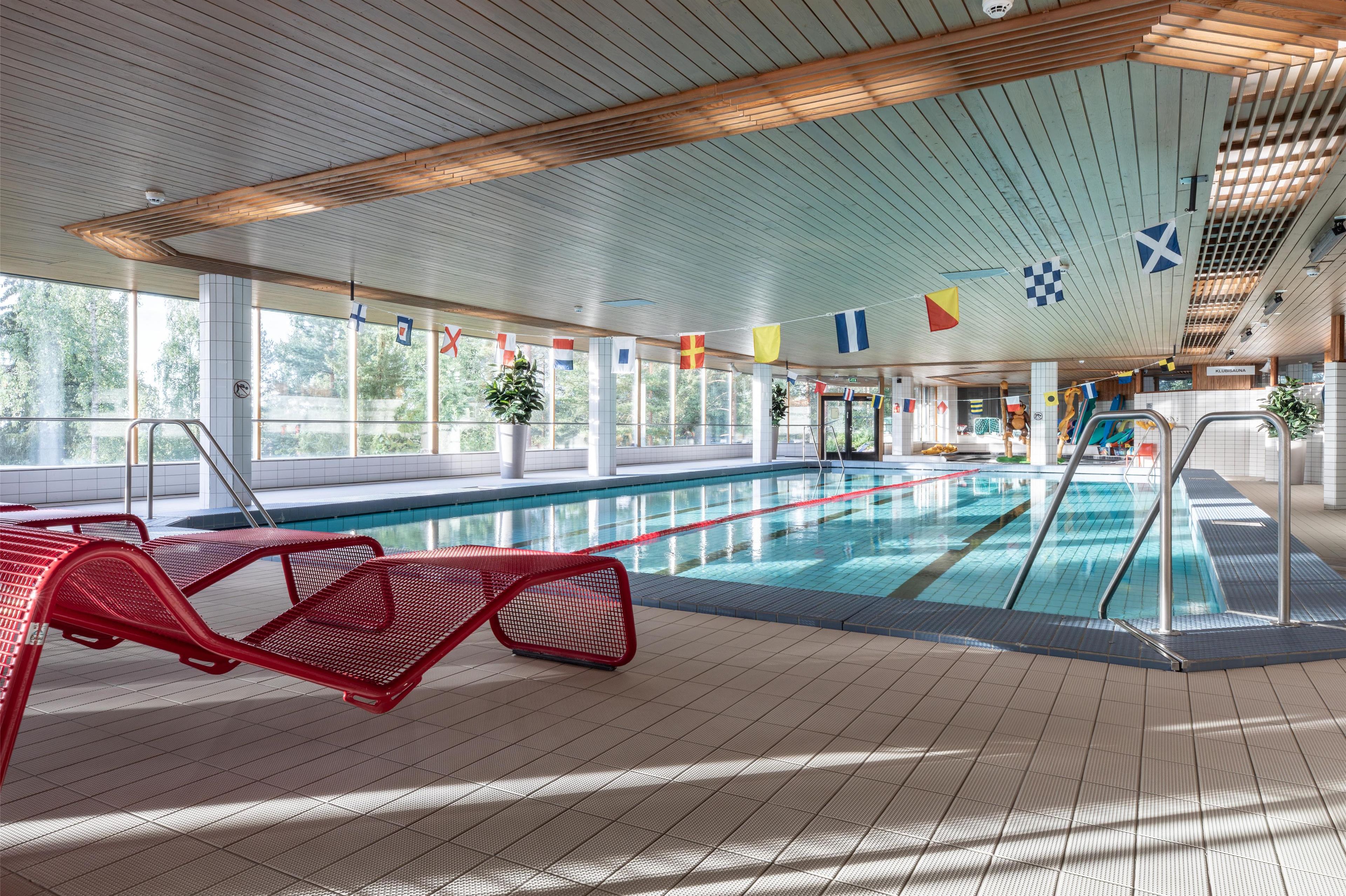 Una piscina interior con suelo de baldosas, tumbonas rojas y banderas colgadas del techo.