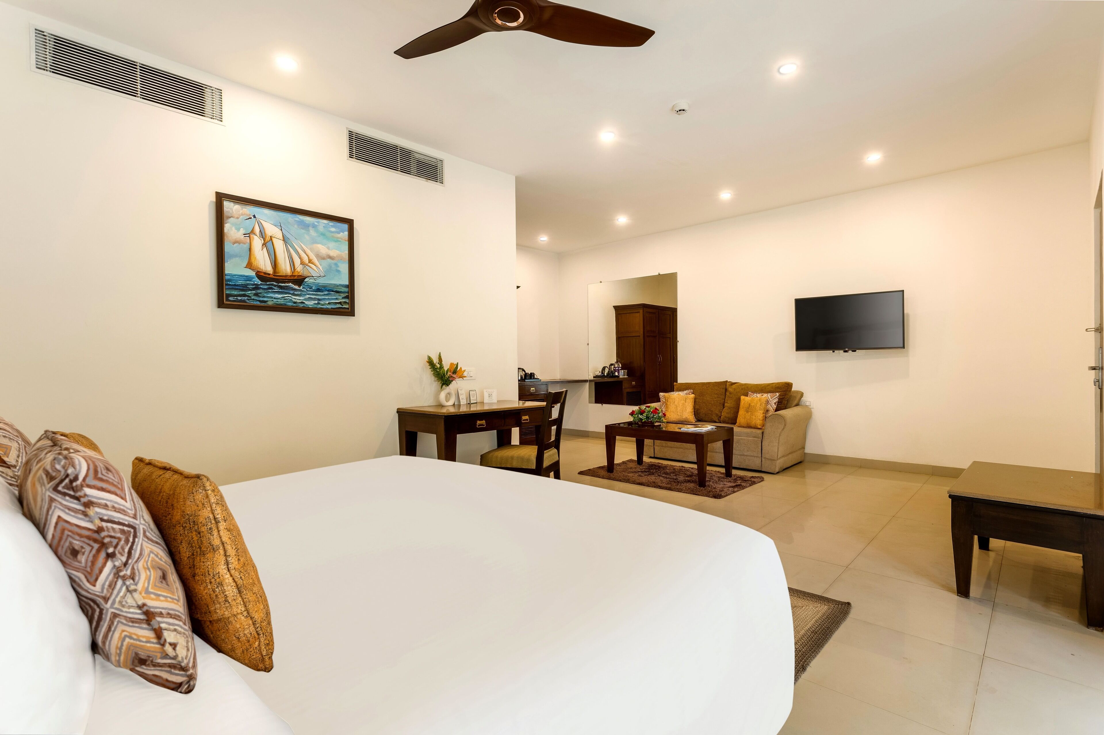 Une chambre d’hôtel avec un grand lit, un bureau avec une chaise, un canapé, un téléviseur fixé au mur et un ventilateur de plafond.