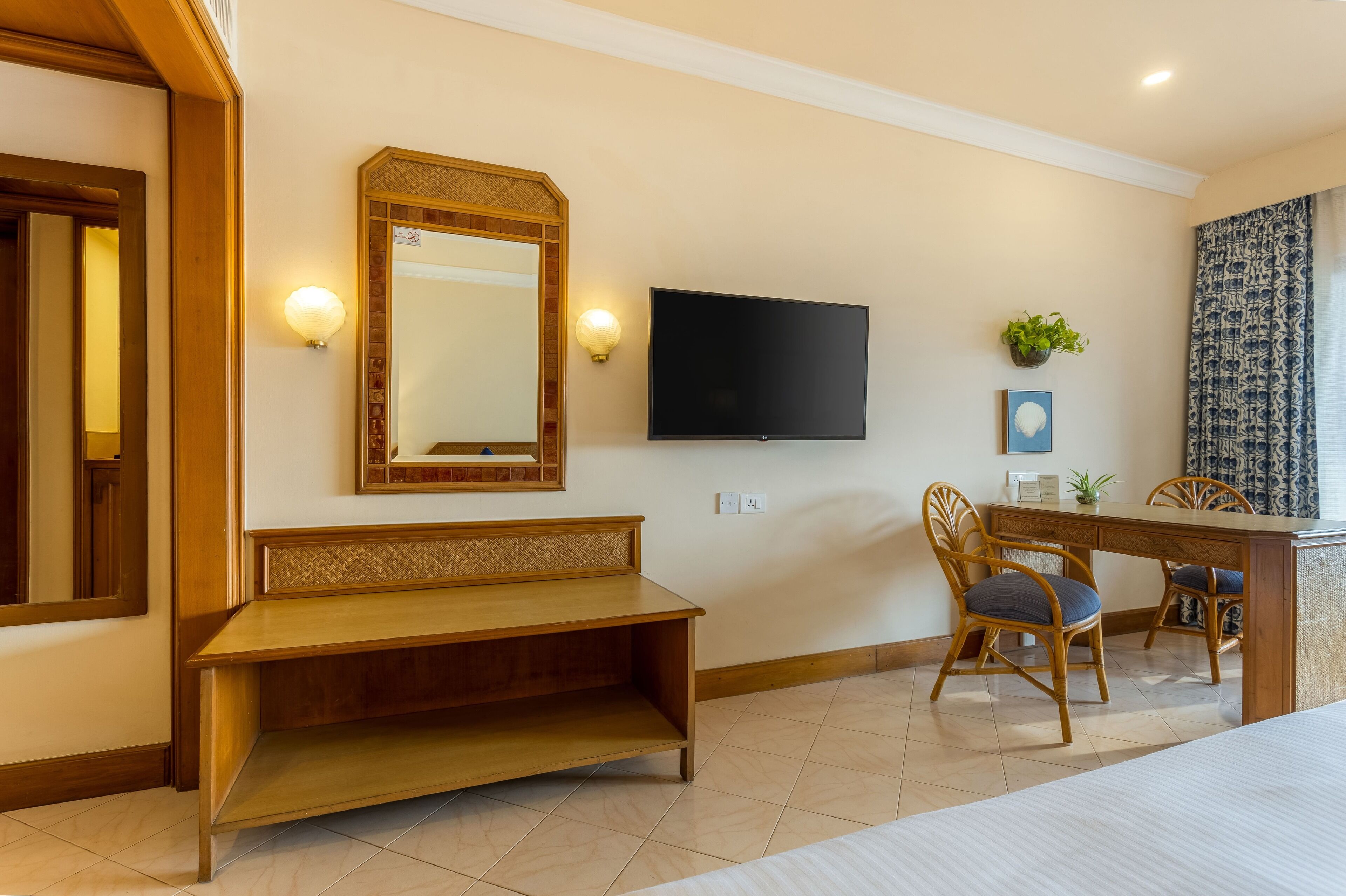 A hotel room with a wooden bench, a TV mounted on the wall, and two wicker chairs.