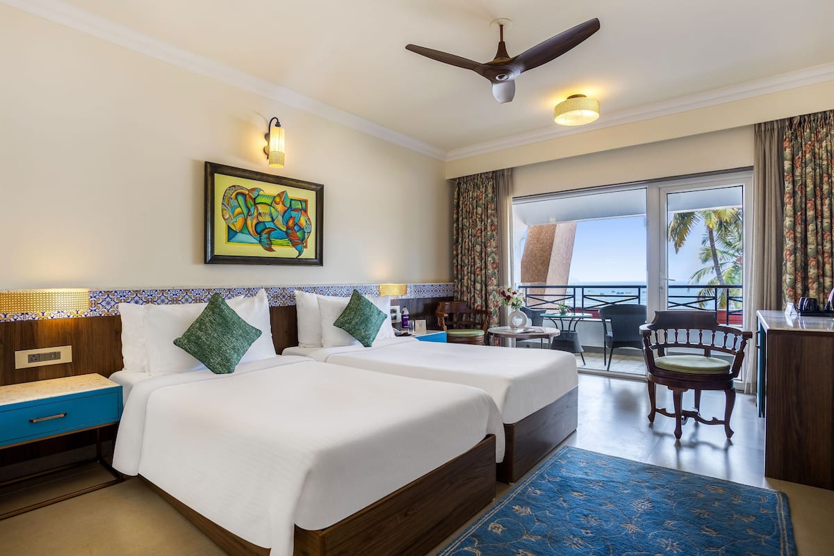 A hotel room with two beds, a ceiling fan, a painting, a balcony with a view, and a desk with a chair.